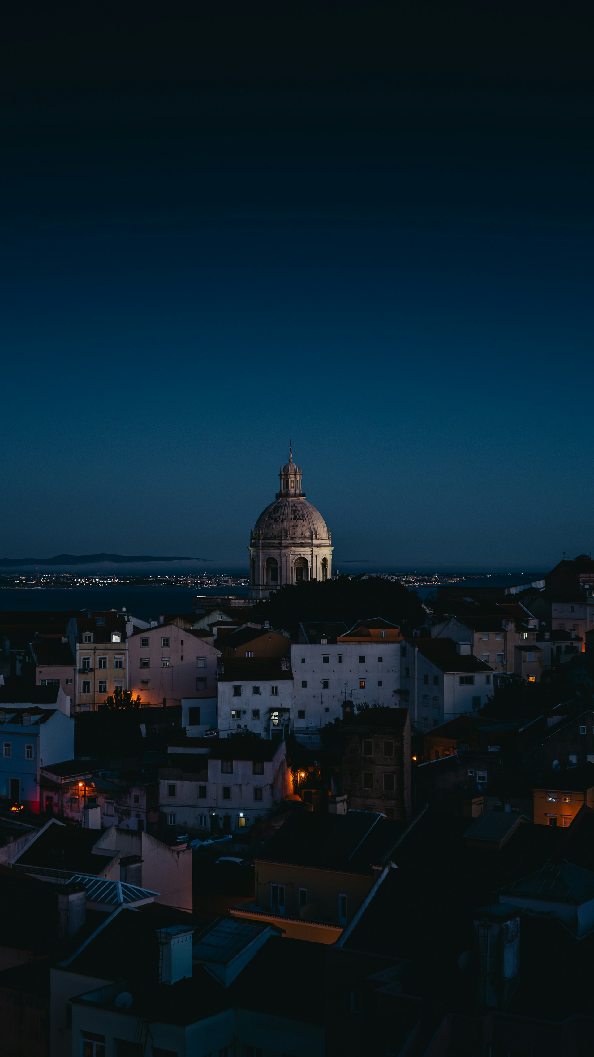 Nighttime cityscape with a large illuminated domed building in the center and surrounding houses under a dark blue sky.