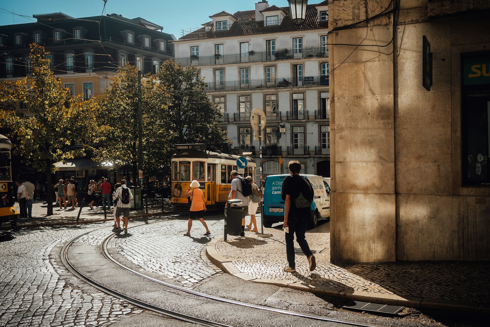 Sunlit urban street scene with people walking near a yellow tram on cobblestone tracks and historic buildings.