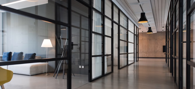 Modern office corridor with glass walls, a gray couch, floor lamp, and black pendant lights.