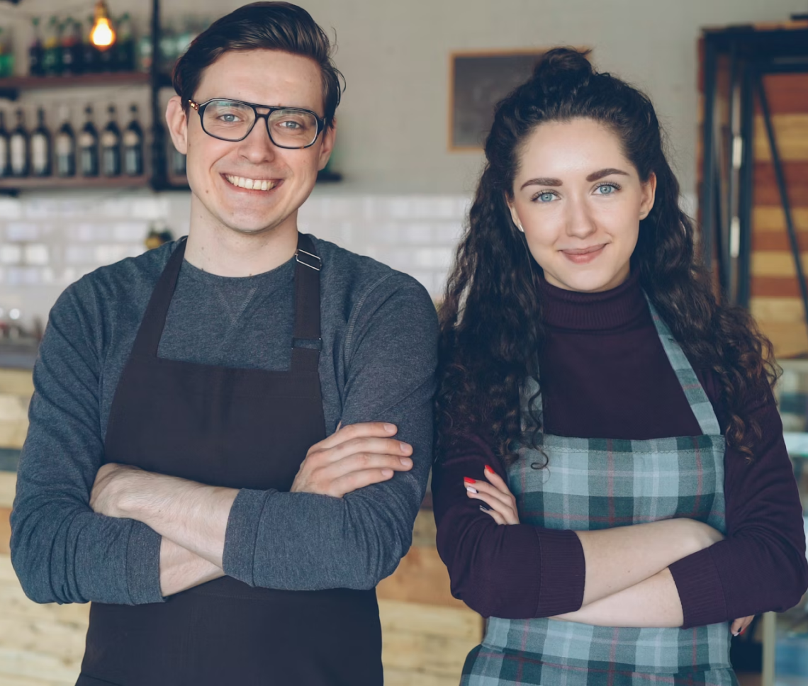 Smiling male and female cafe workers standing with arms crossed in a coffee shop.