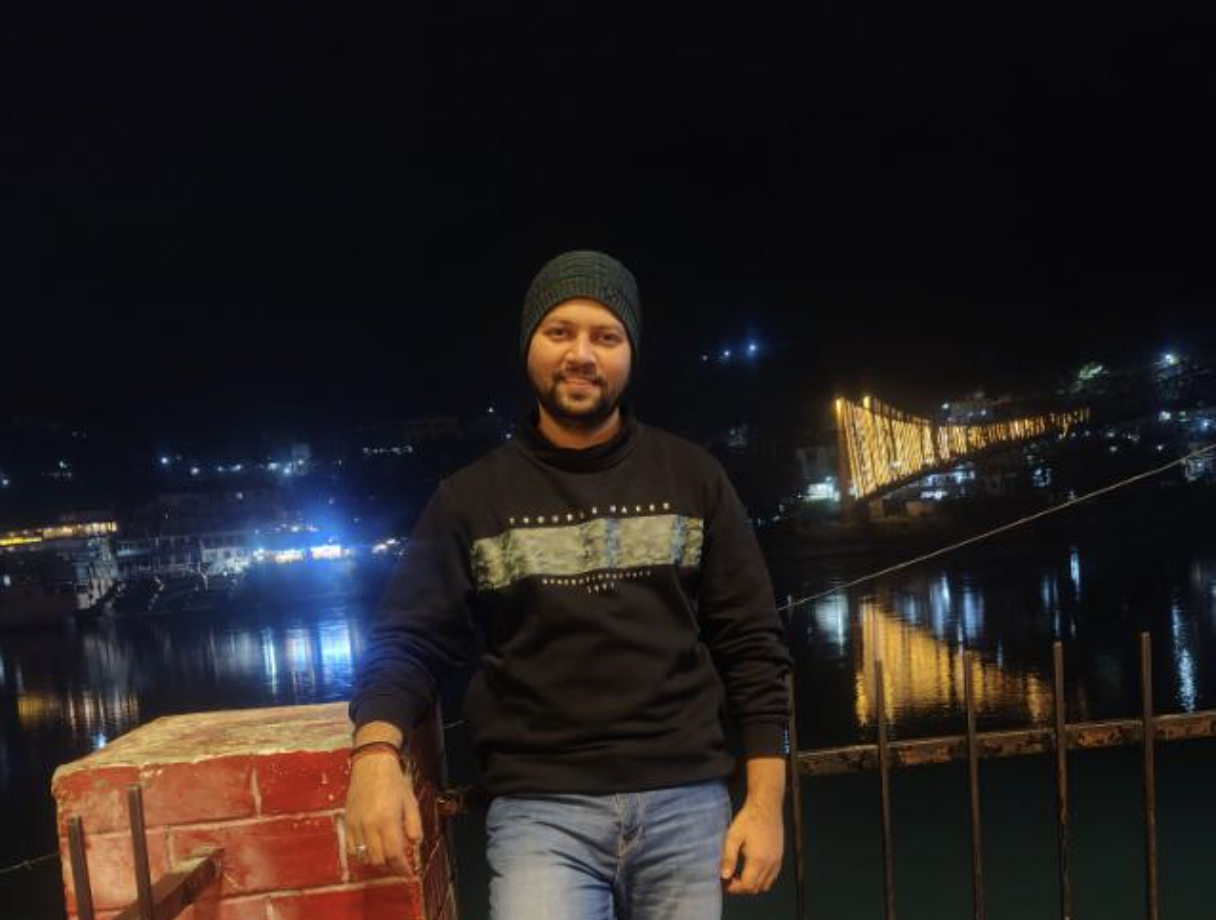 Man wearing a black beanie and sweater leaning on a red brick wall at night with a brightly lit bridge and water reflections in the background.
