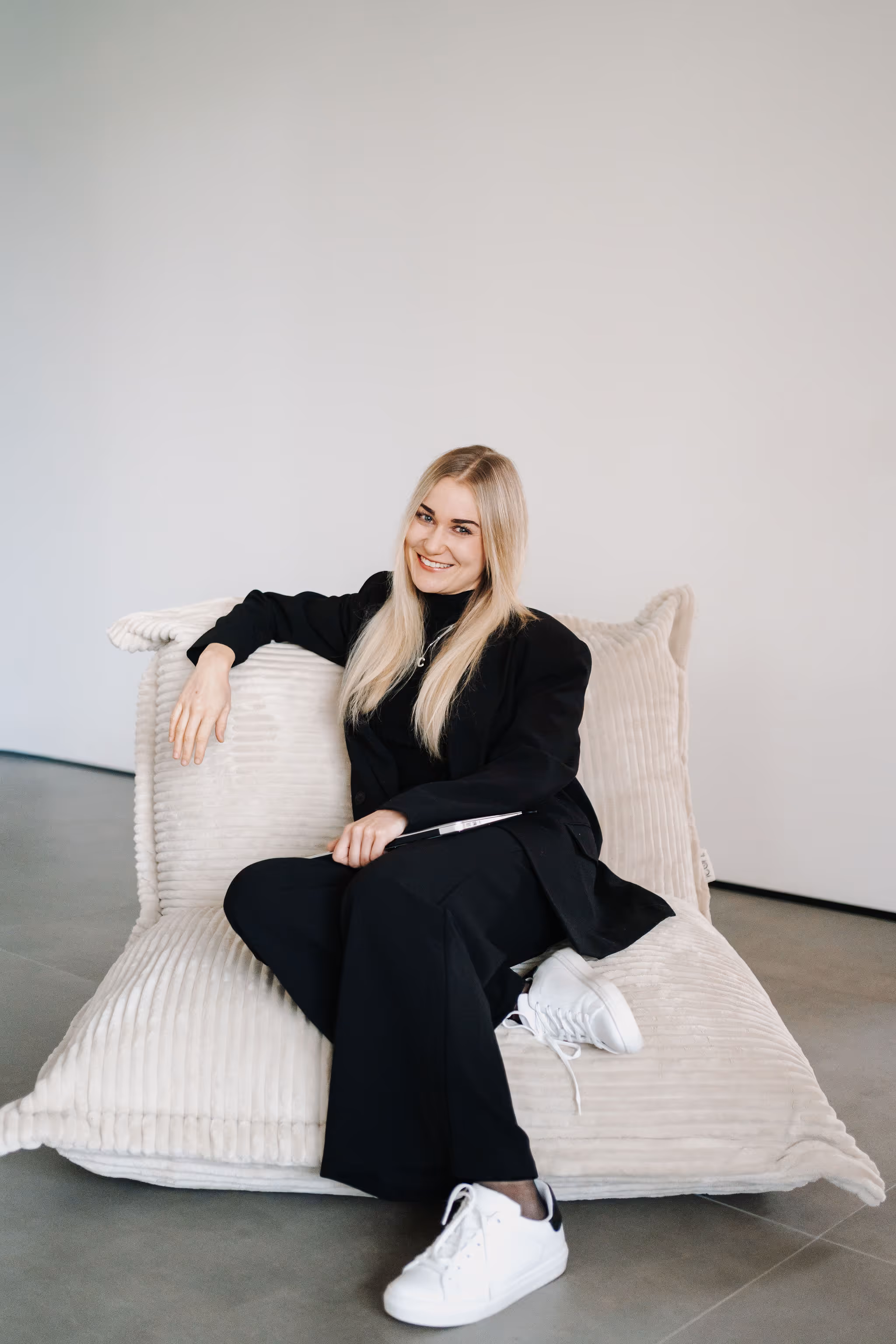 Junge Frau mit langen blonden Haaren in schwarzem Outfit und weißen Sneakers sitzt lächelnd auf einem großen, beigen, kuscheligen Bodenkissen.
