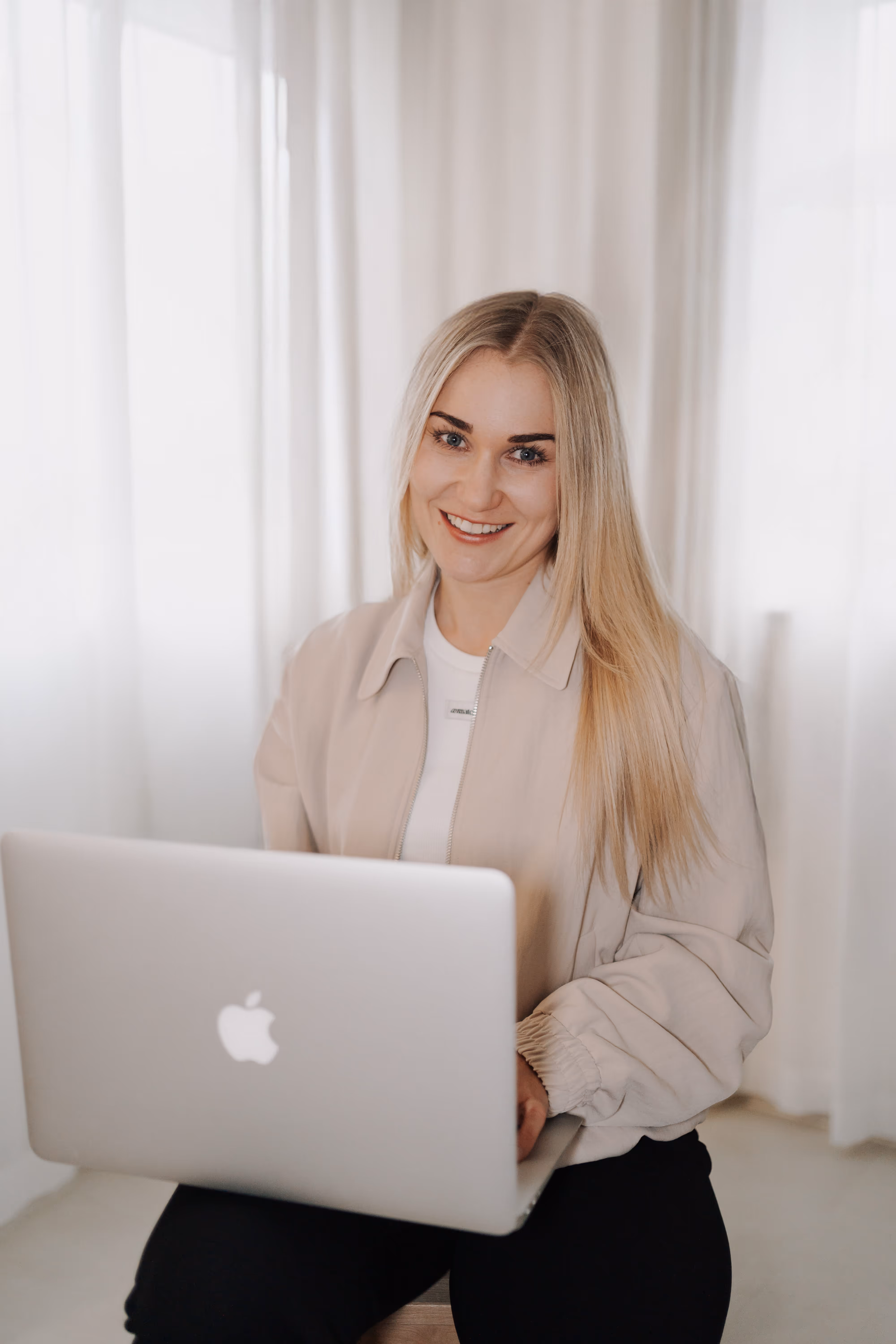 Lächelnde blonde Frau, die vor einem Laptop sitzt und in einem hellen Raum arbeitet.