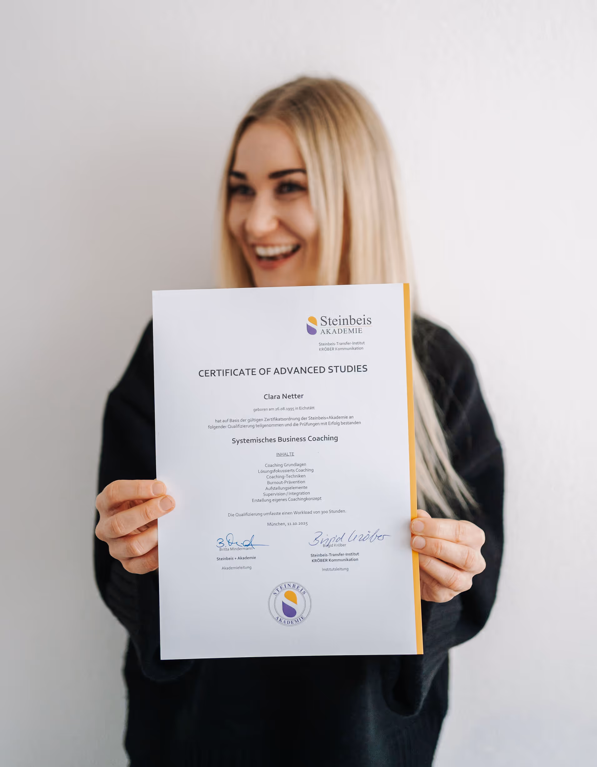Frau mit blonden Haaren hält ein Zertifikat für Systemisches Business Coaching der Steinbeis Akademie in die Kamera und lächelt dabei.
