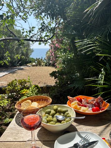 Tisch mit einem Glas Roséwein, einer Schale mit grünen Oliven, einem Korb mit Brot und einem Teller mit verschiedenen Aufschnitt- und Gemüsesorten, vor einer mediterranen Gartenlandschaft mit Blick auf das Meer.