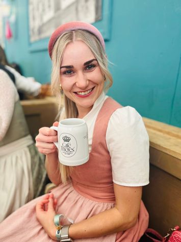 Lächelnde junge Frau in traditioneller Tracht mit rosa Dirndl und Barett hält einen weißen Bierkrug in einem Gastraum.