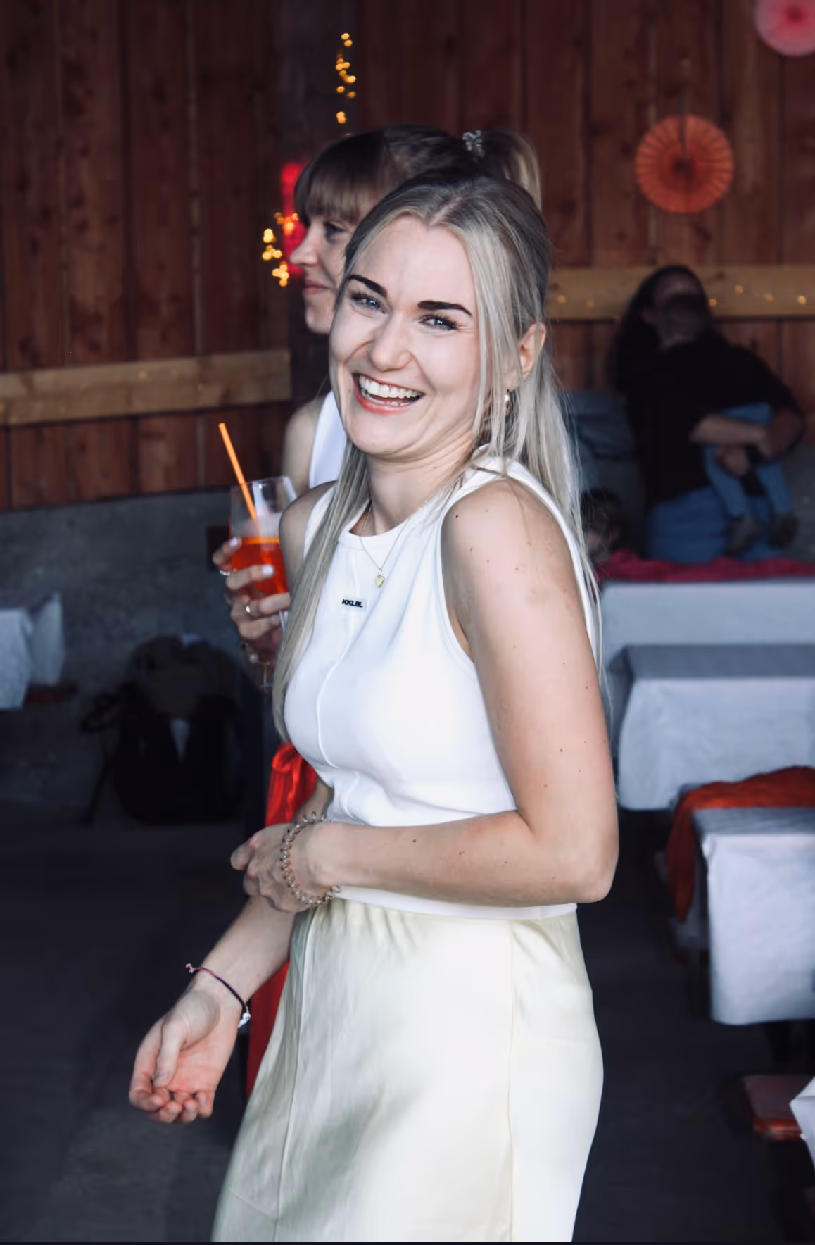 Lächelnde blonde Frau mit weißem Oberteil und hellem Rock auf einer Party in einem Raum mit Holzwänden.