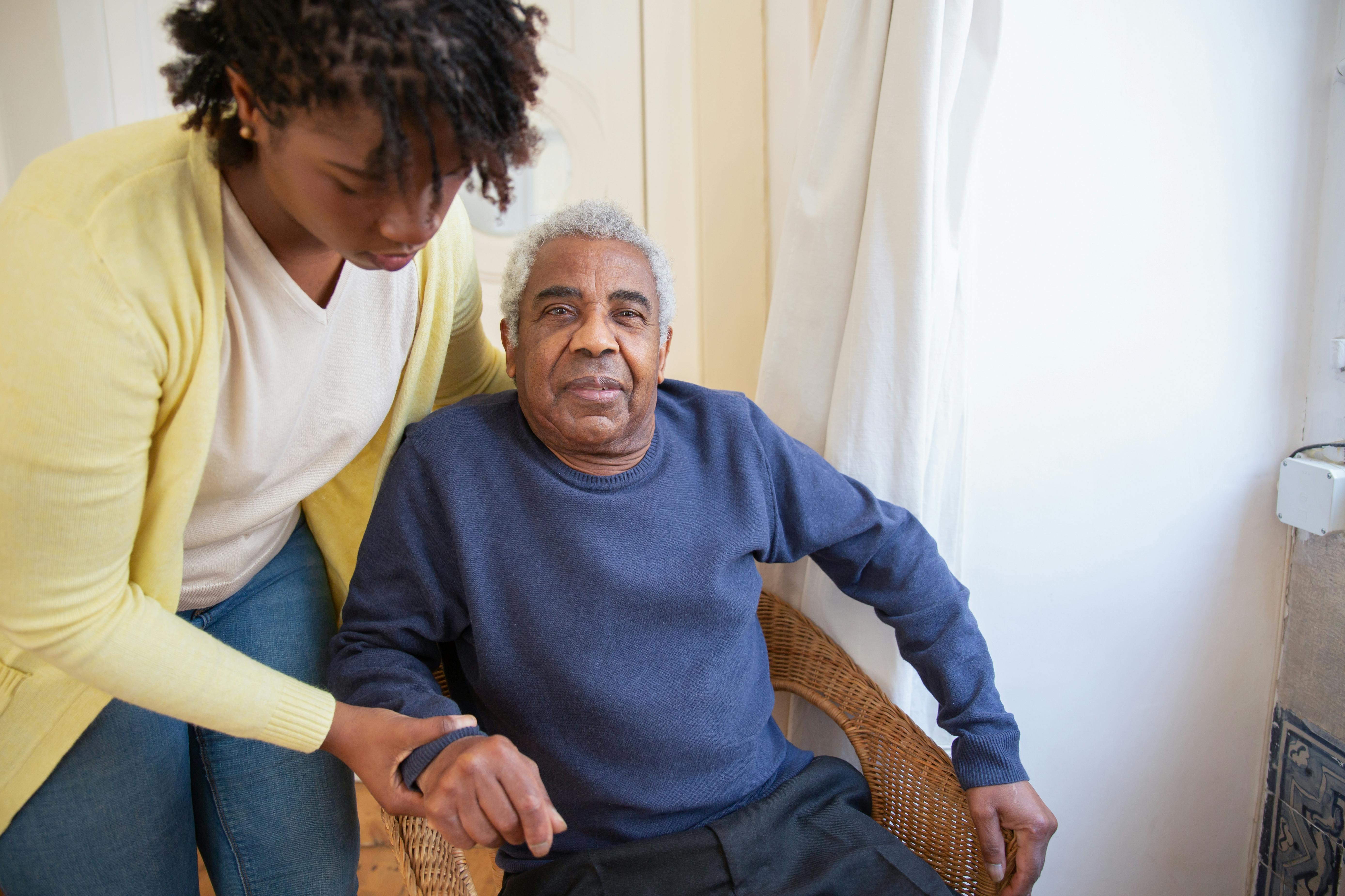Caregiver supporting elderly man sitting in a wicker chair indoors.