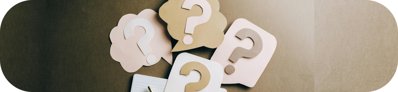 Several paper speech bubbles with question marks resting on a brown surface next to a pen and notebook.