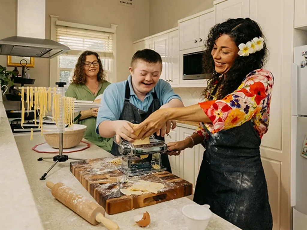 Three people in a kitchen making fresh pasta with a pasta machine, smiling and enjoying the activity.