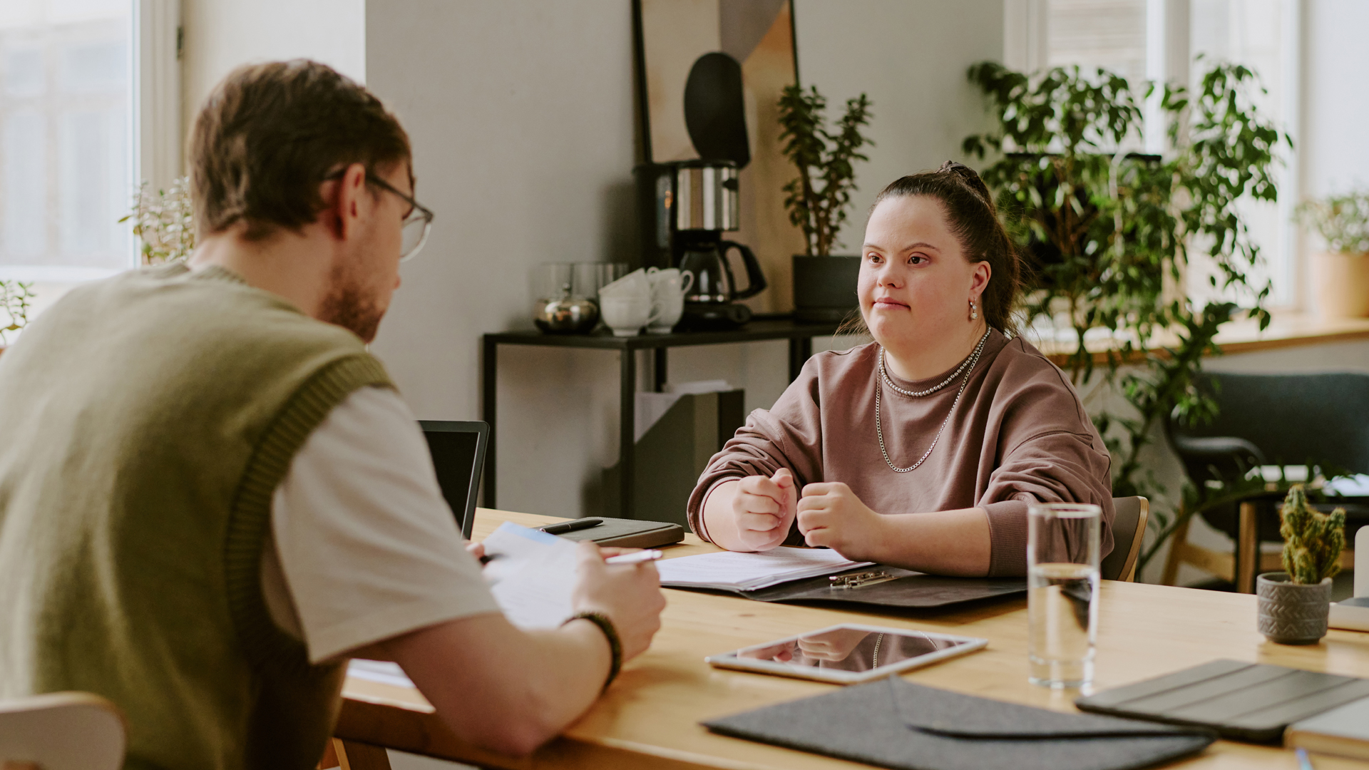 A woman with Down syndrome listens attentively across a wooden table to a man reviewing documents during a meeting in a cozy office space.