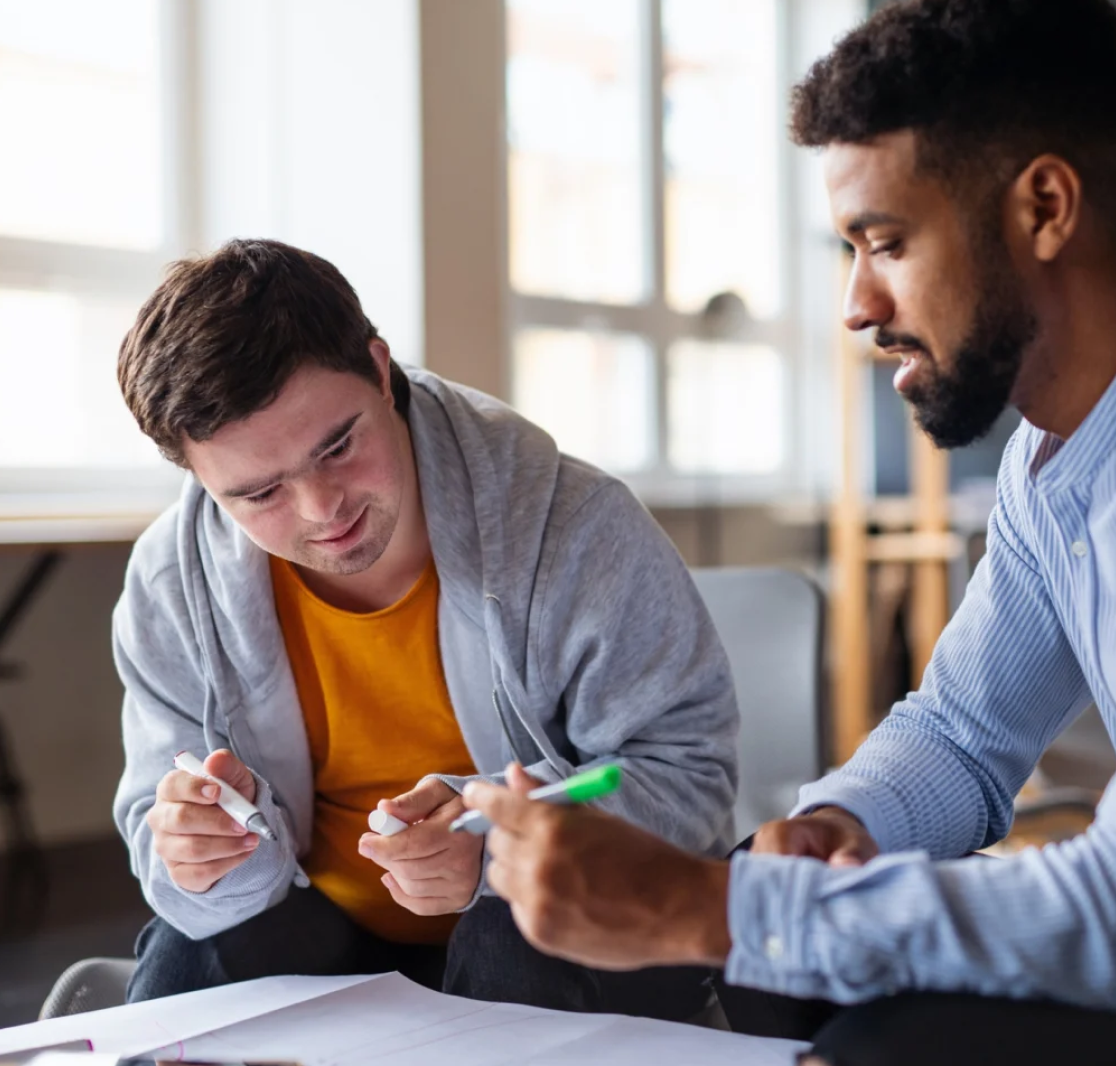 Two men, one with Down syndrome, collaborating and discussing notes with pens in hand in a bright office setting.
