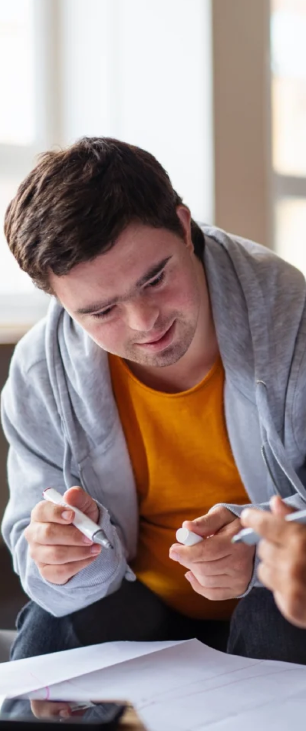 Young man in a gray hoodie and orange shirt holding a whiteboard marker, engaged in a discussion with another person pointing with a green marker over papers.