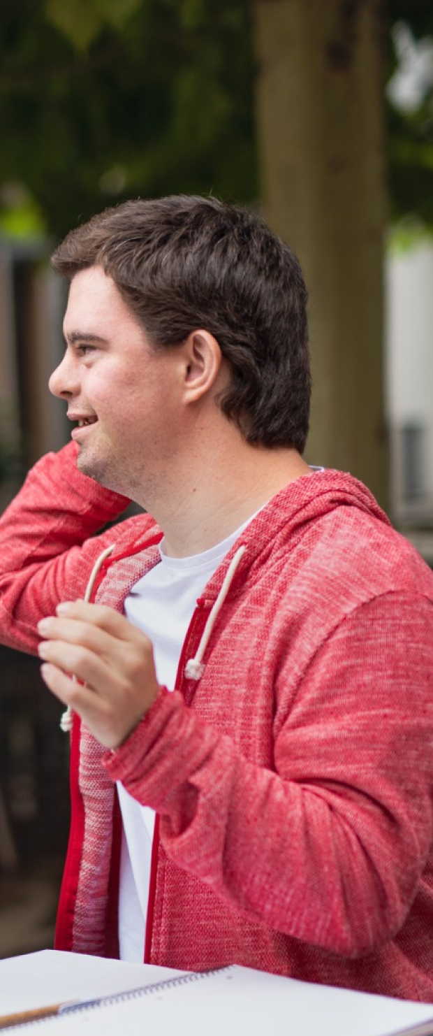 Young man in a red hoodie and white shirt smiling and adjusting his hair outdoors.