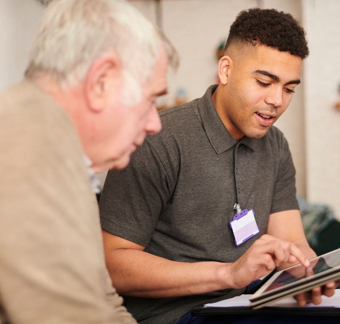 Young man with name badge showing tablet screen to an elderly man in a helping or teaching context.