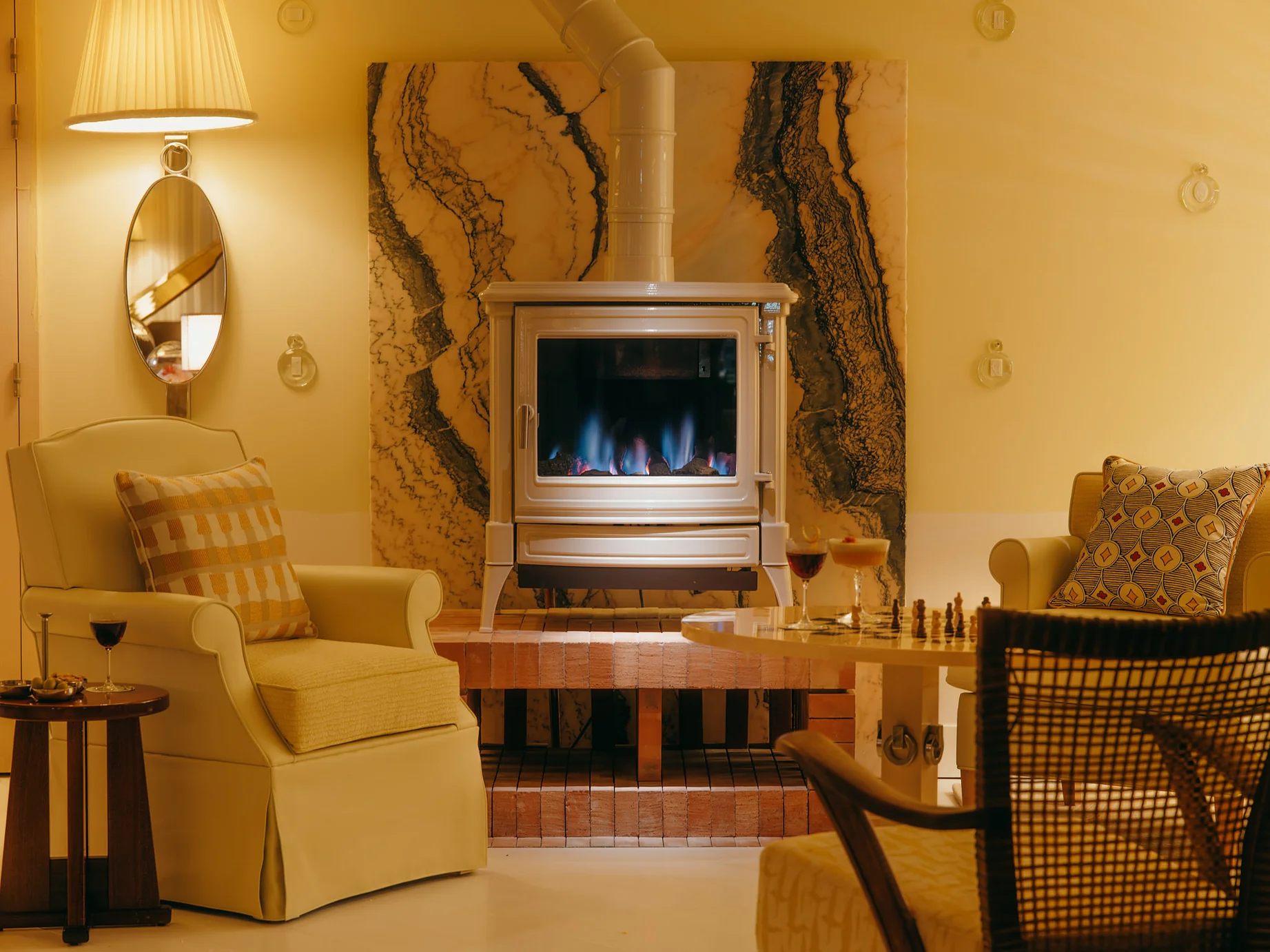 Coin du bar de la Villa Colette avec poêle à bois allumé sur fond de marbre veiné, fauteuils crème, verre de vin rouge et jeu d'échecs