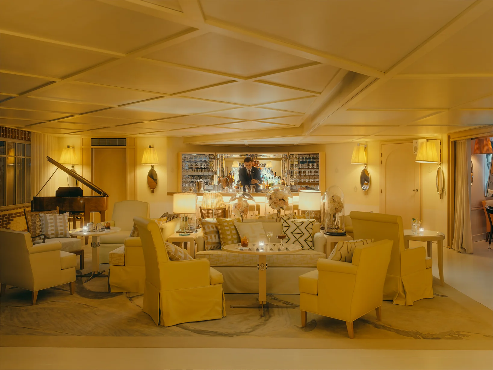 Bar et salon de la Villa Colette en soirée avec piano à queue, fauteuils jaunes, comptoir de bar éclairé, fleurs sous cloche et plafond à caissons