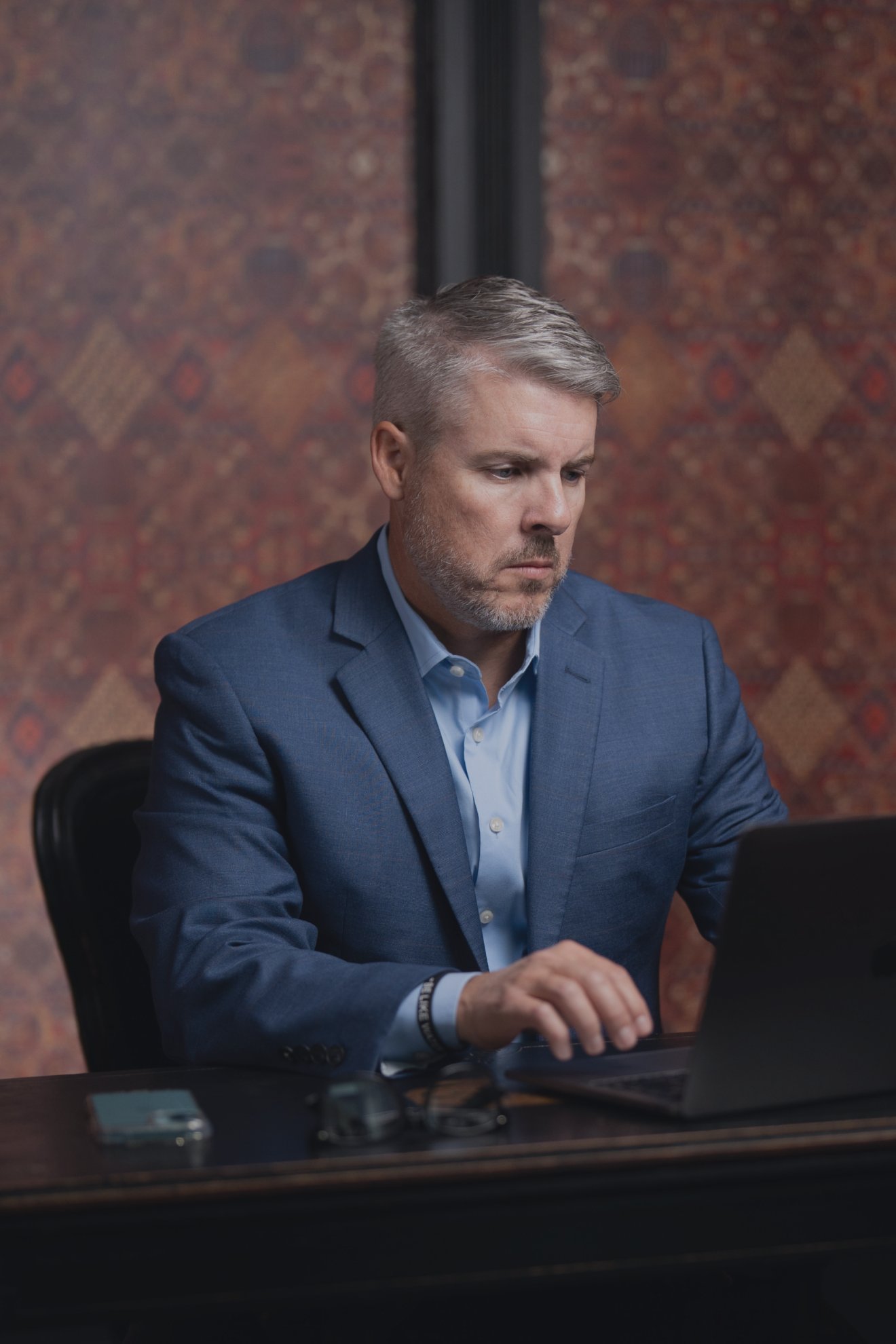 Middle-aged man in a blue suit focused on typing on a laptop at a desk with a phone and glasses.