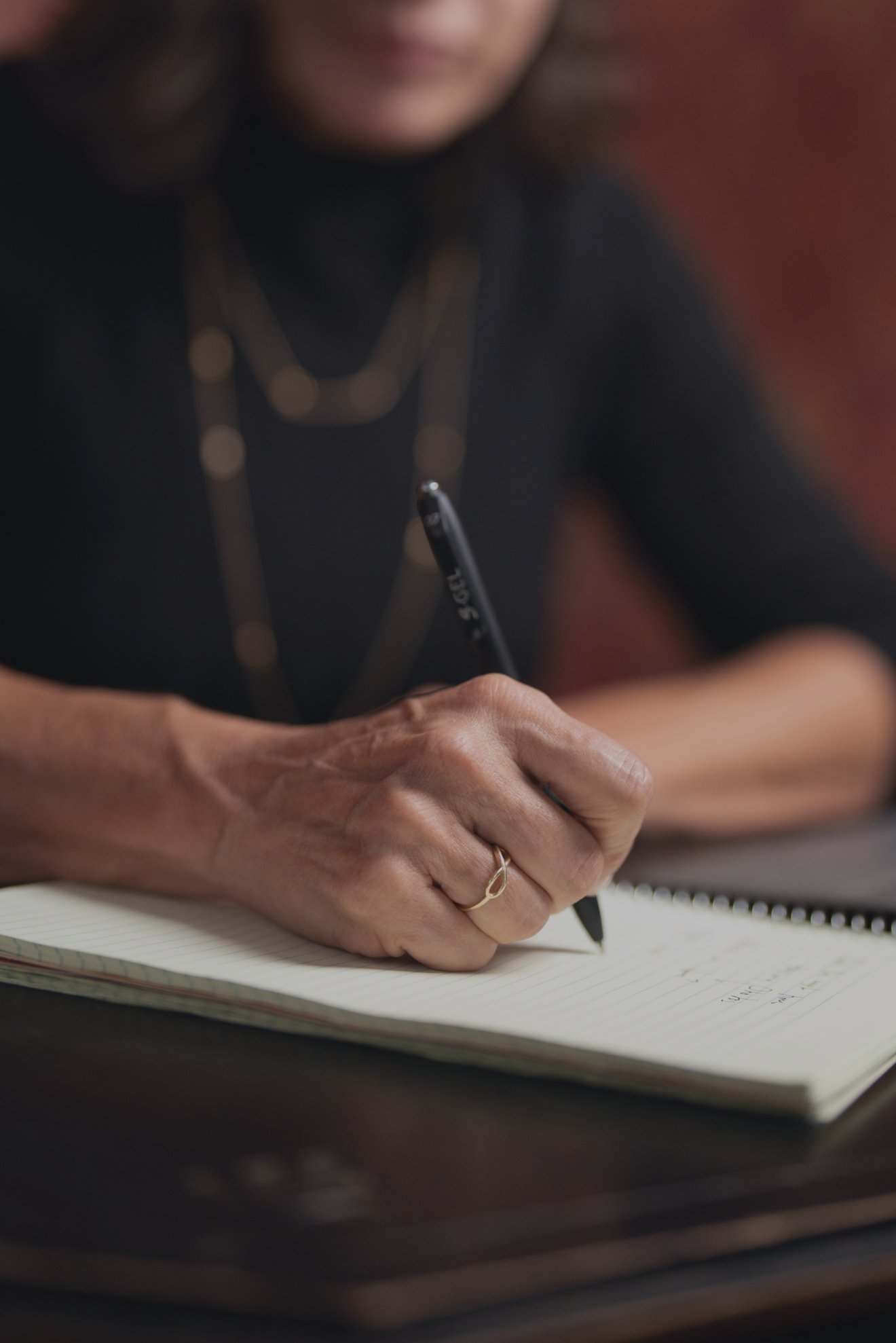 Close-up of a person wearing a gold ring writing with a black pen on a lined notebook.