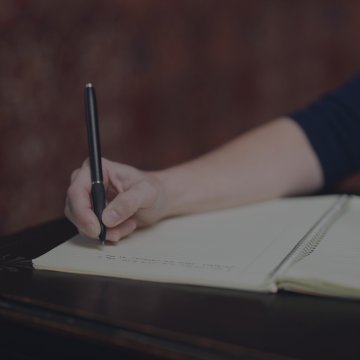 Person writing in a spiral notebook with a pen on a dark table.