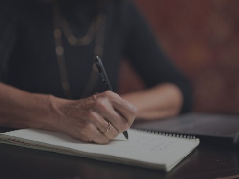 Person writing on a spiral notebook with a pen at a desk.
