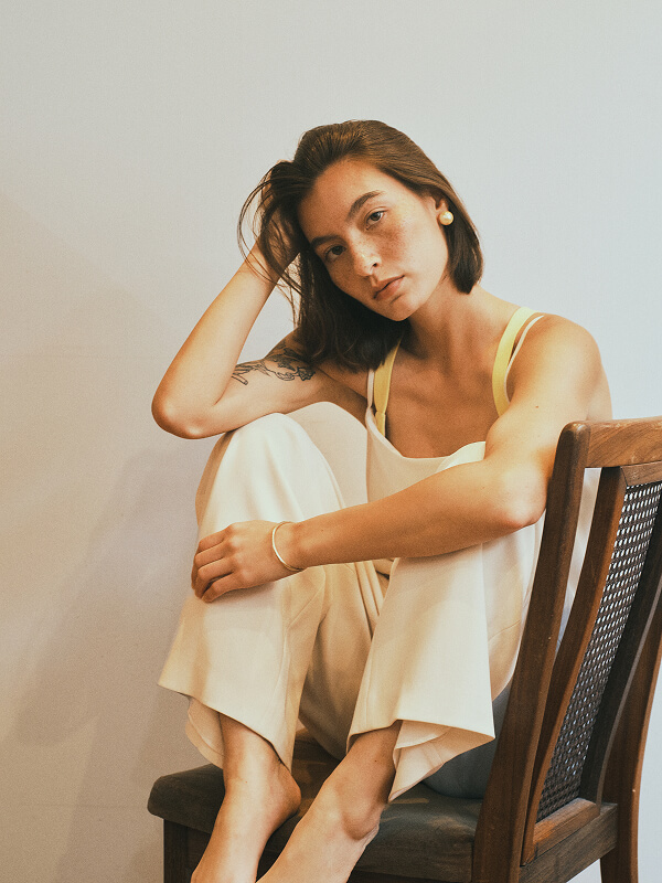 Woman with short brown hair, wearing a sleeveless yellow top and cream pants, sitting barefoot on a wooden chair against a plain light wall.