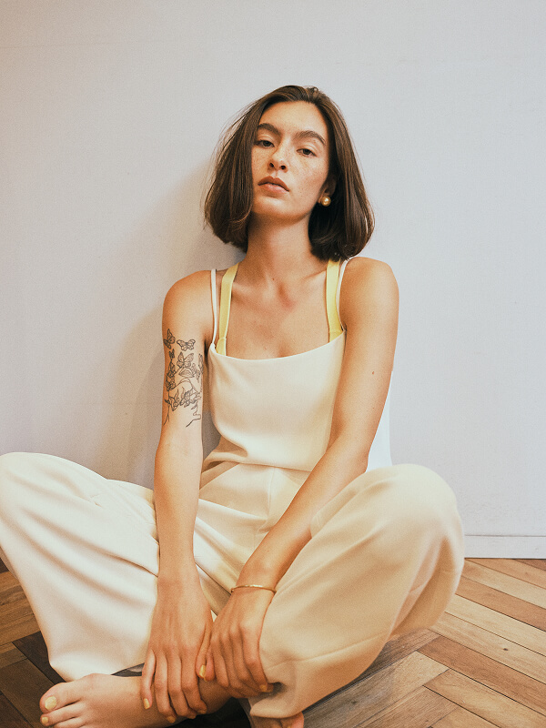 Young woman with bob haircut and floral tattoo on left arm wearing cream overalls sitting cross-legged on wooden floor against a plain white wall.