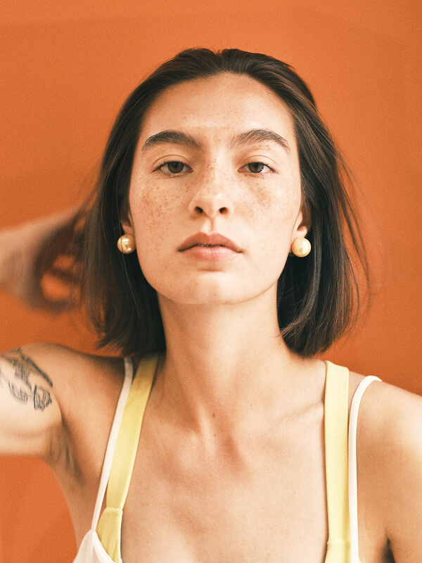 Portrait of a woman with shoulder-length brown hair, wearing pearl earrings and a yellow-strapped top against an orange background.