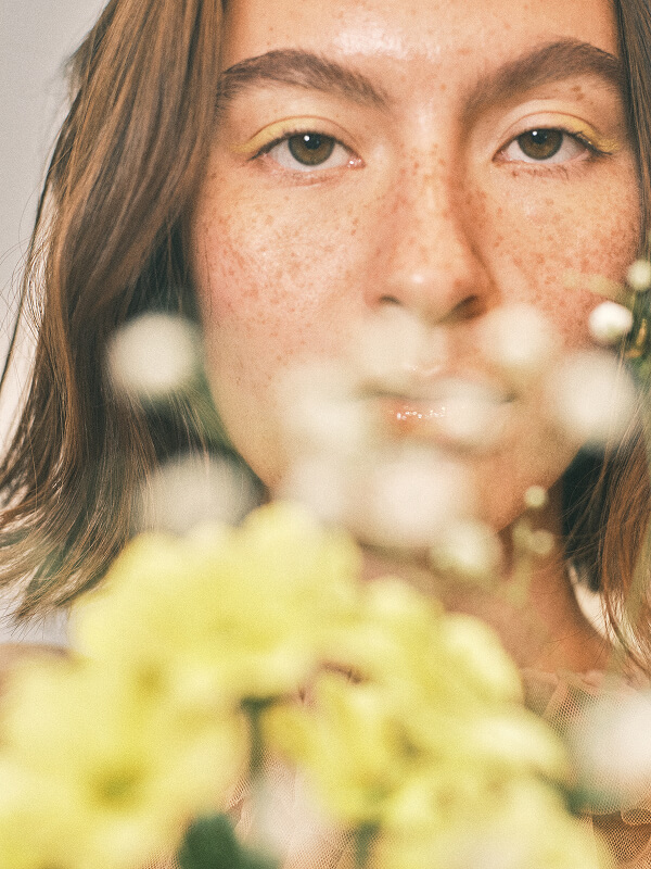 Close-up of a freckled woman's face with yellow eyeshadow, partially obscured by out-of-focus yellow and white flowers.