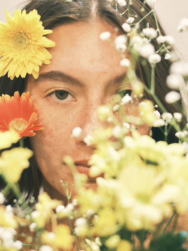 Close-up of a young woman with freckles surrounded by yellow and orange flowers, some flowers partially obscuring her face.