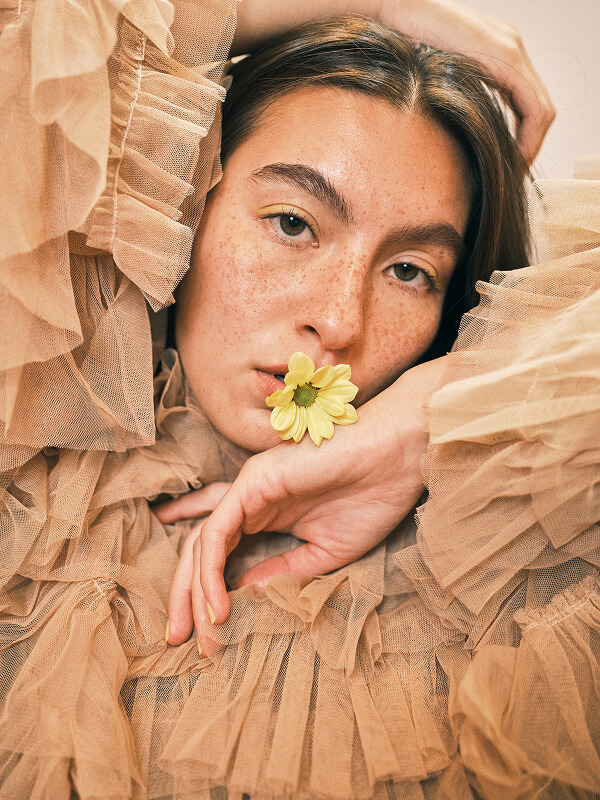 Close-up of a woman with freckles wearing a beige ruffled mesh top, holding a small yellow flower near her lips.