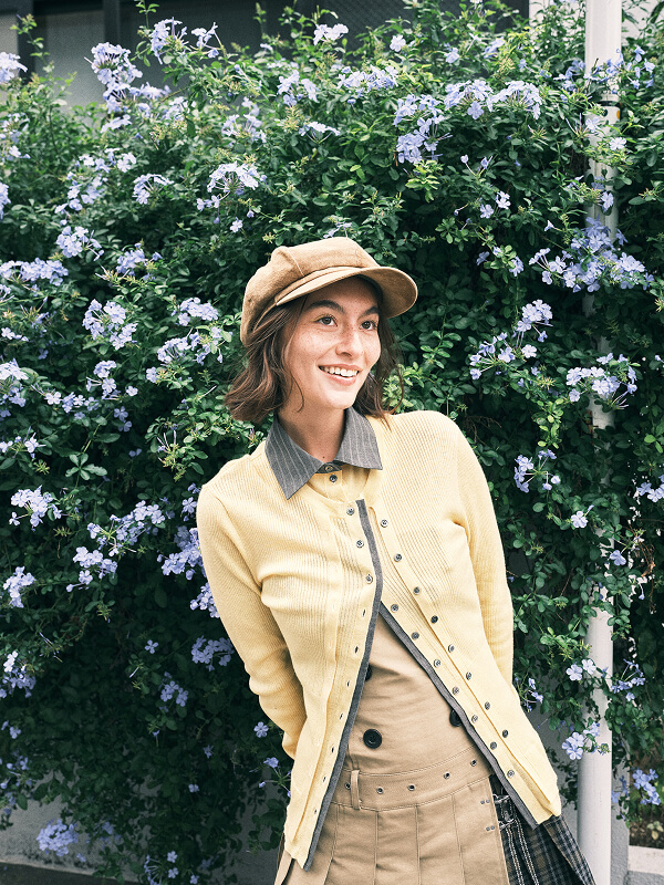 Smiling woman in a yellow cardigan and beige cap standing in front of a lush green bush with small purple flowers.