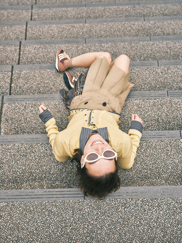 Smiling woman wearing white sunglasses and a yellow coat lying on outdoor stone steps.