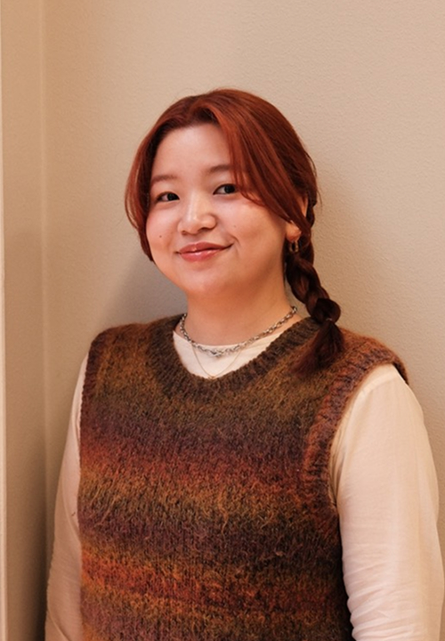 Woman with shoulder-length auburn hair wearing black glasses and a gray sweater, looking to the side.