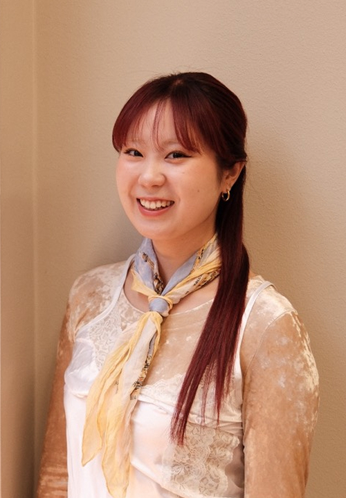 Young woman with long, wavy, dark pink hair wearing a red sweater, smiling over her shoulder indoors.