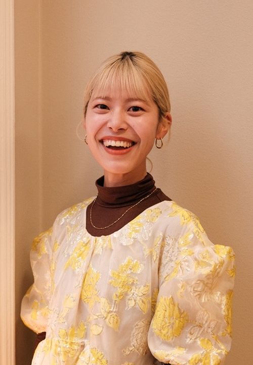 Smiling woman with blonde hair wearing a yellow and white floral dress over a brown turtleneck standing against a beige wall.