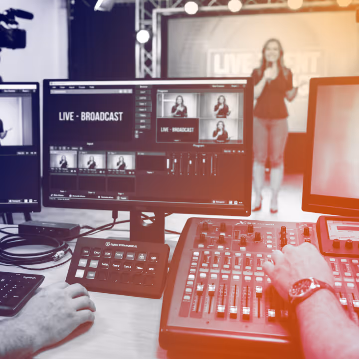 Person controlling audio mixer and video switcher during live broadcast with presenter on stage.