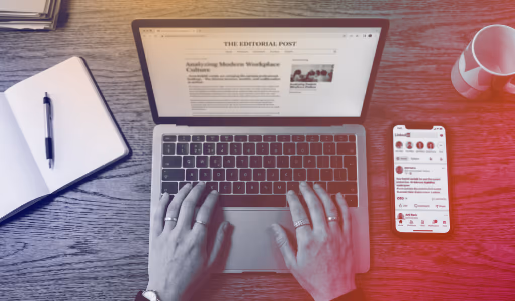 Hands typing on a laptop with an article on screen, beside a notebook, smartphone, and coffee mug.