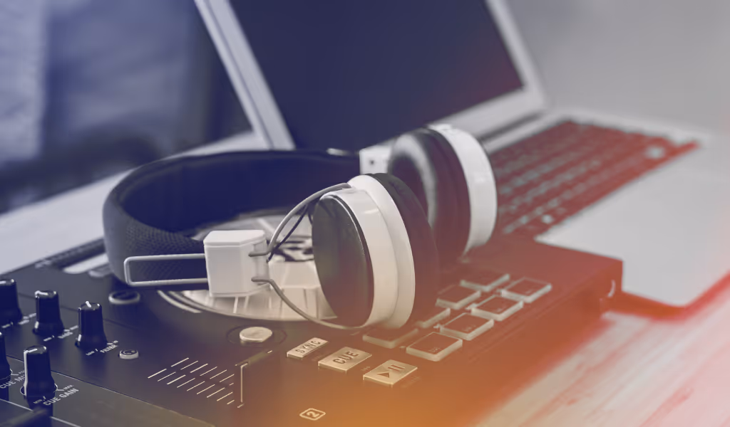 Headphones resting on a DJ mixer with a laptop in the background on a wooden surface.