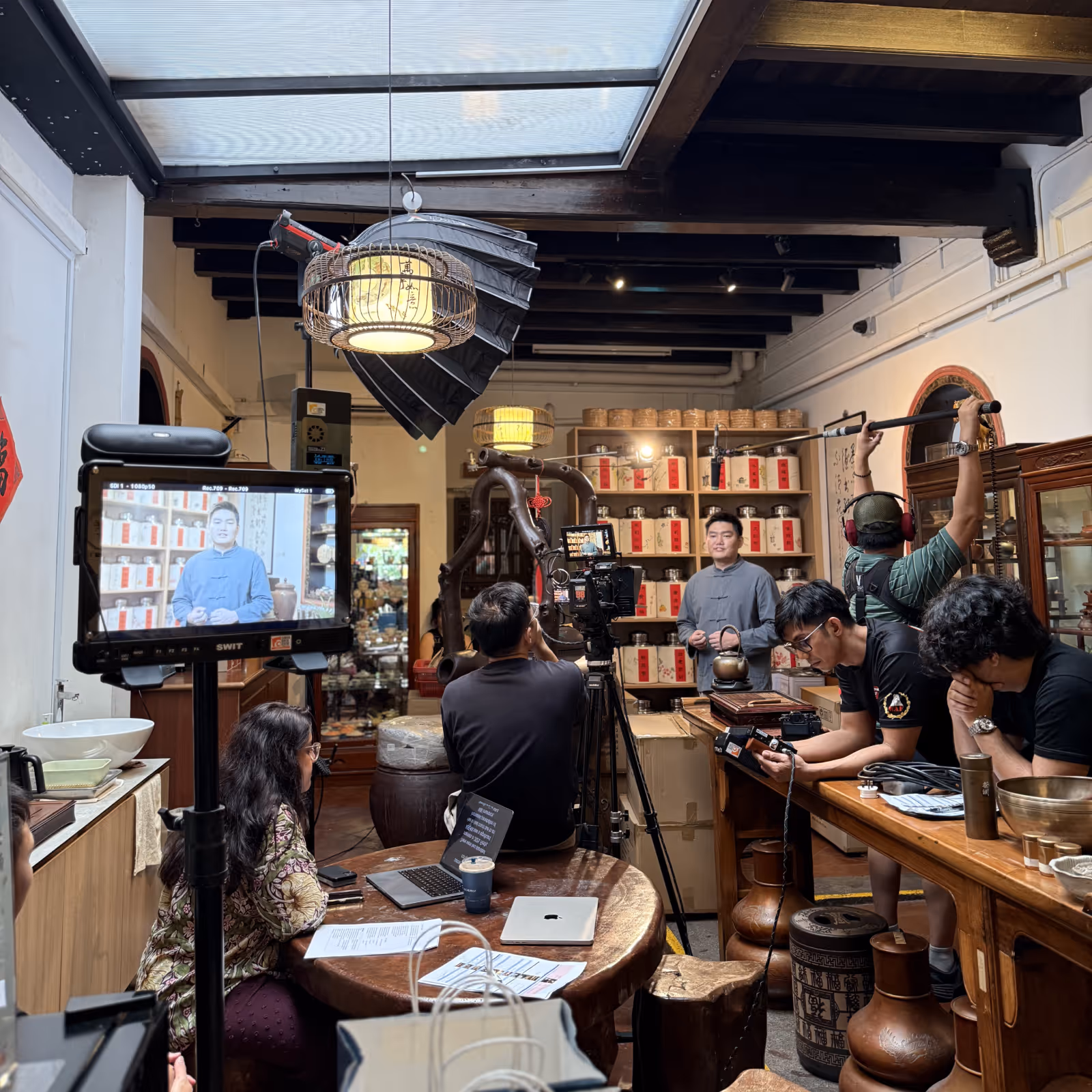 Film crew recording a man speaking in a traditional tea shop with lighting and sound equipment set up.