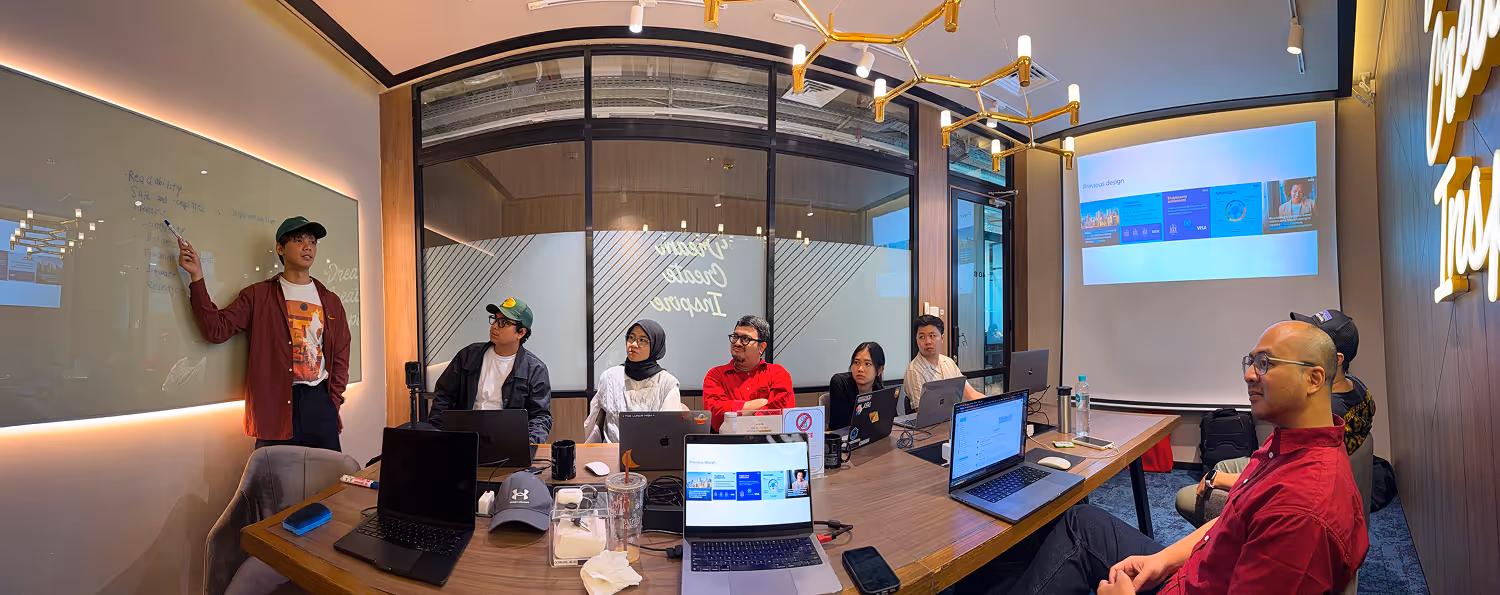 A group of eight people in a modern conference room with laptops, one person presenting at a whiteboard.