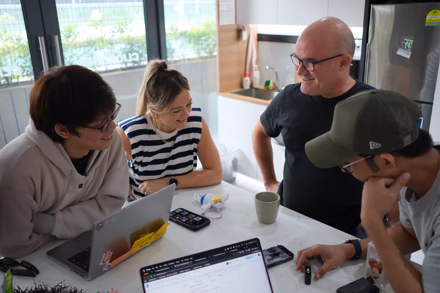 Four people smiling and interacting around a kitchen table with laptops, phones, and a coffee mug.