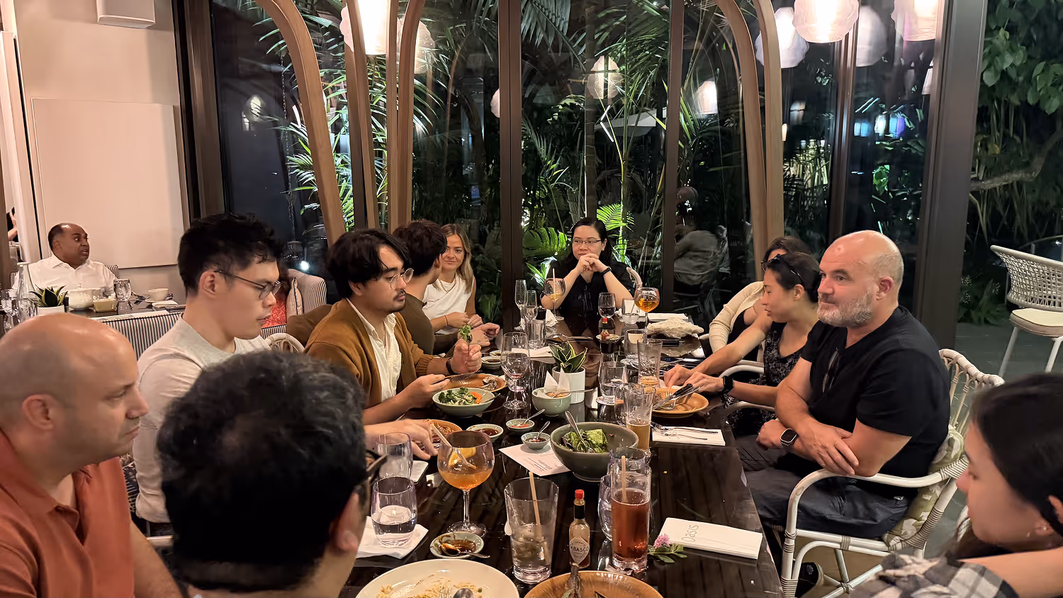 Group of people seated around a long table with food and drinks in a dining room at night.