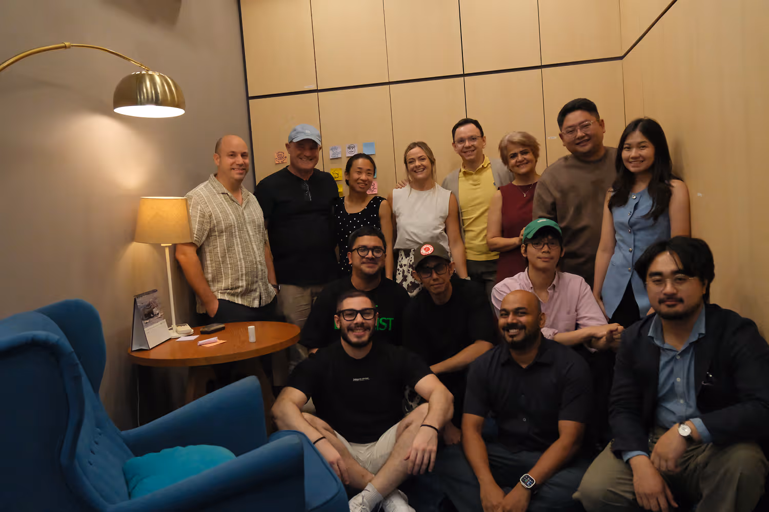 Group of 13 diverse people smiling and posing in a cozy room with blue chairs and wall lamp.