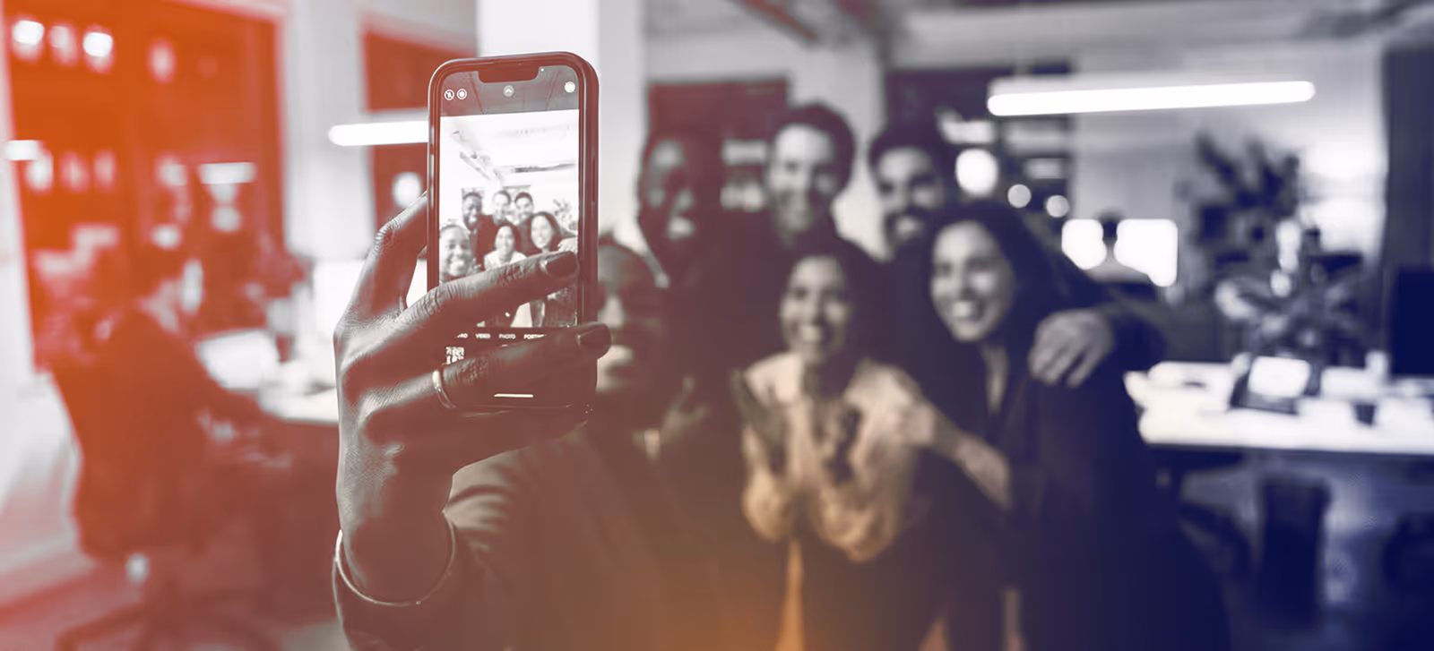 A group of coworkers smiling and posing for a selfie taken with a smartphone in an office.