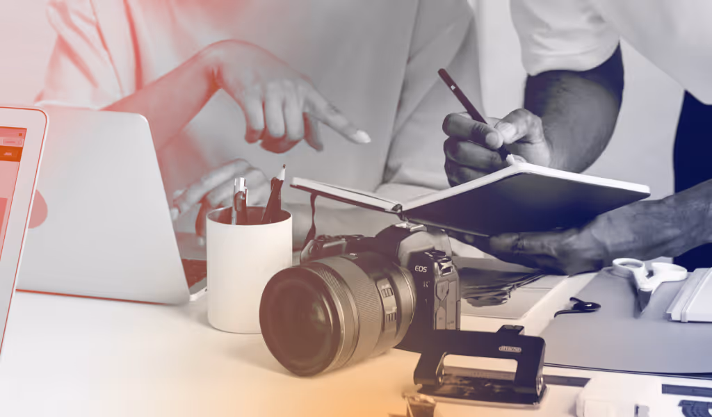 Hands pointing at and writing in a notebook near a camera, laptop, and desk supplies.