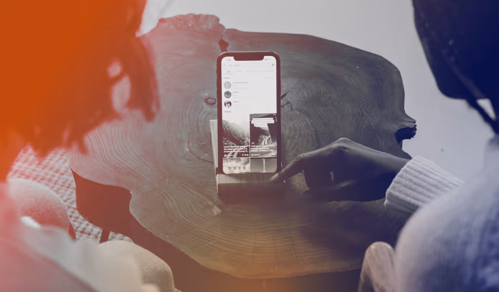 Two people sitting by a wooden table, one pointing to a smartphone screen displaying a social media app.