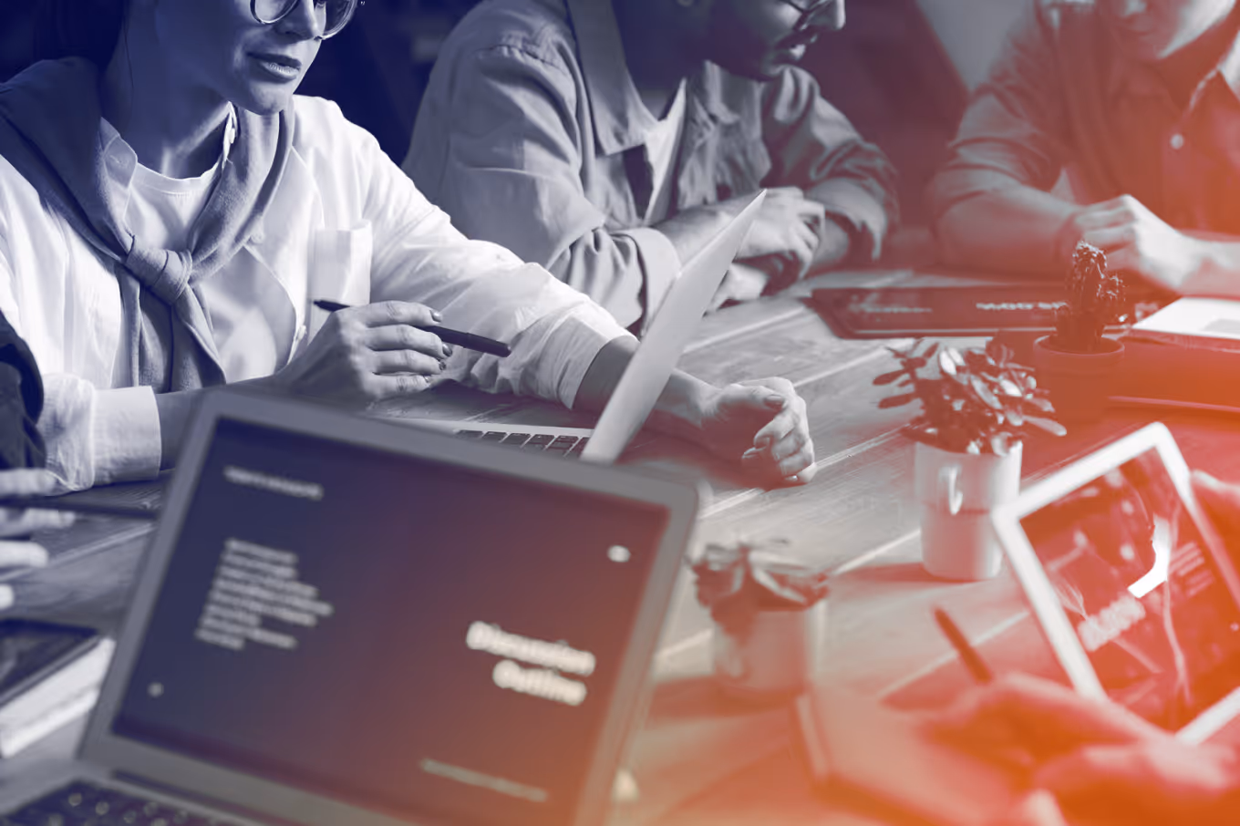 People working together at a table with laptops, tablets, notebooks, and small potted plants.