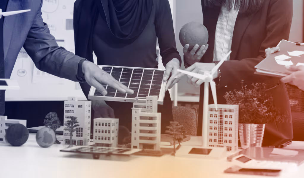People examining solar panel and wind turbine models on a table with miniature buildings and trees.