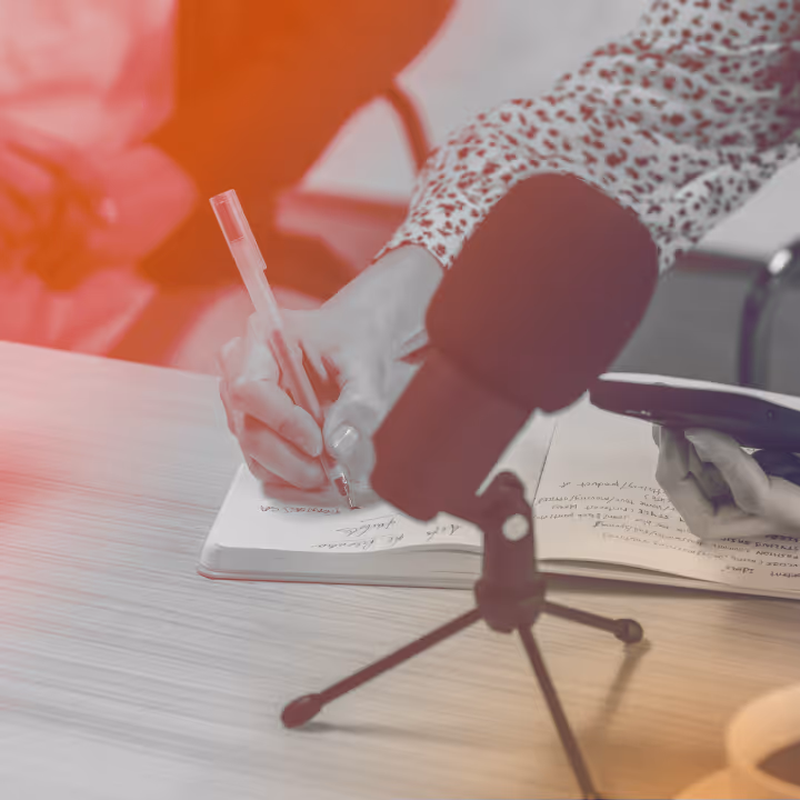 Person writing notes in a notebook with a microphone and another hand holding a device.